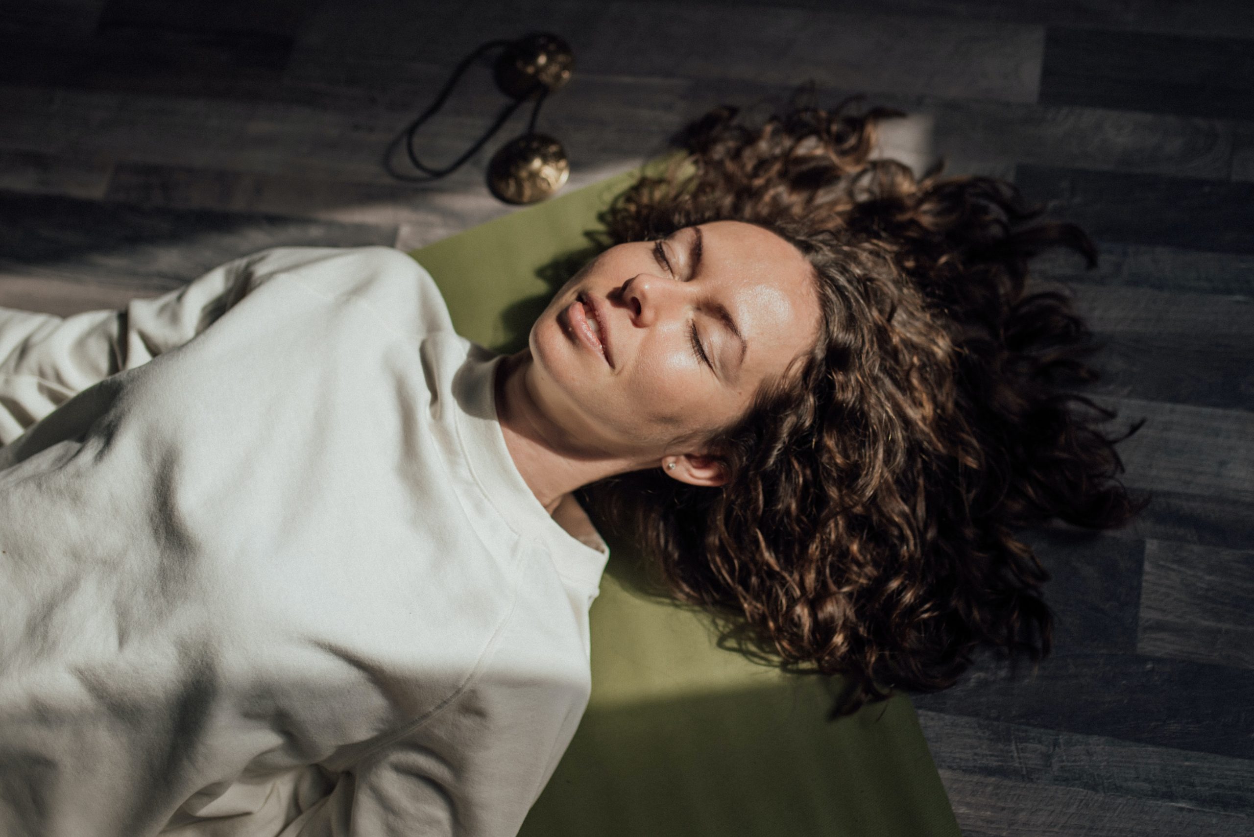 A white woman with short and curly hair is lying down smiling with her eyes shut. She is on a green yoga mat and wears a white sweater.,